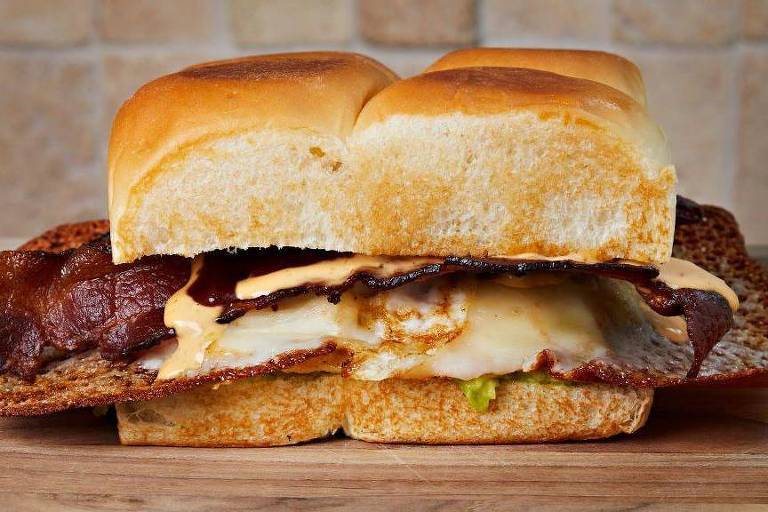 Um suculento sanduíche de bacon e queijo derretido repousa sobre uma tábua de madeira, com pães dourados e macios abraçando as camadas de carne e queijo.