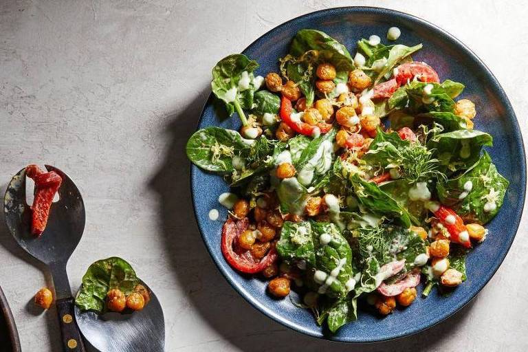Um prato azul escuro apresenta uma salada repleta de folhas verdes, grão-de-bico dourado e tomates vermelhos vibrantes. Ao lado, uma colher de salada com alguns ingredientes sugere que a refeição está prestes a ser servida