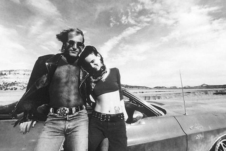 Imagem em preto e branco do filme. Um rapaz e uma mulher jovem estão encostados em um carro estacionado numa paisagem desértica do interior dos EUA