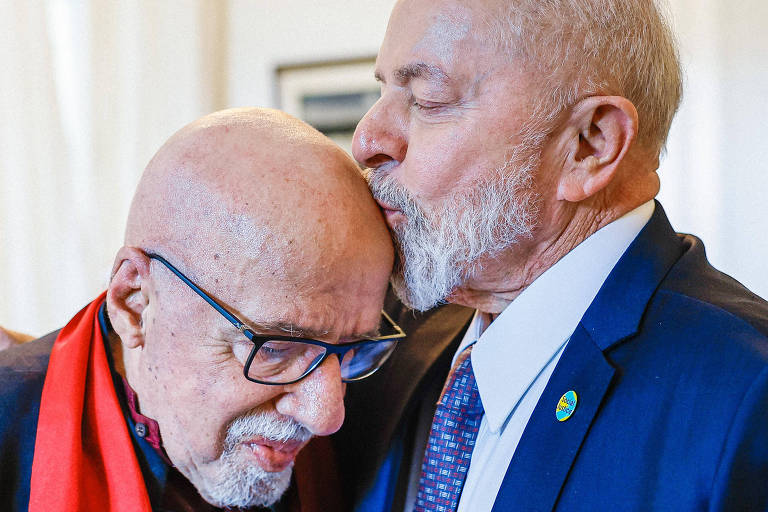 Lula, um homem branco com barba branca e vestindo um terno azul com uma lapela adornada com uma fita verde e amarela, beija a cabeça de Paulo Coelho, um homem branco, careca, que usa óculos e um lenço vermelho