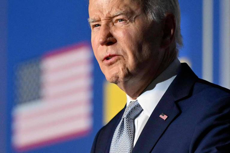 Joe Biden aparece em primeiro plano com uma expressão séria, enquanto ao fundo desfocado, as cores da bandeira dos Estados Unidos podem ser vistas