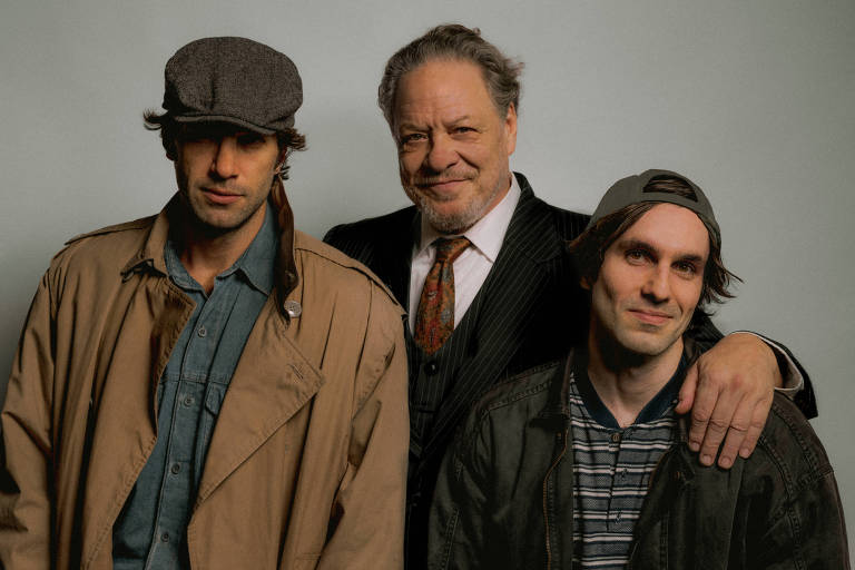 Três homens brancos posam juntos, o homem à esquerda usa uma boina e uma jaqueta sobreposta a uma camisa, enquanto o homem do meio está com um terno e gravata, e o homem à direita veste uma jaqueta sobre uma camiseta listrada