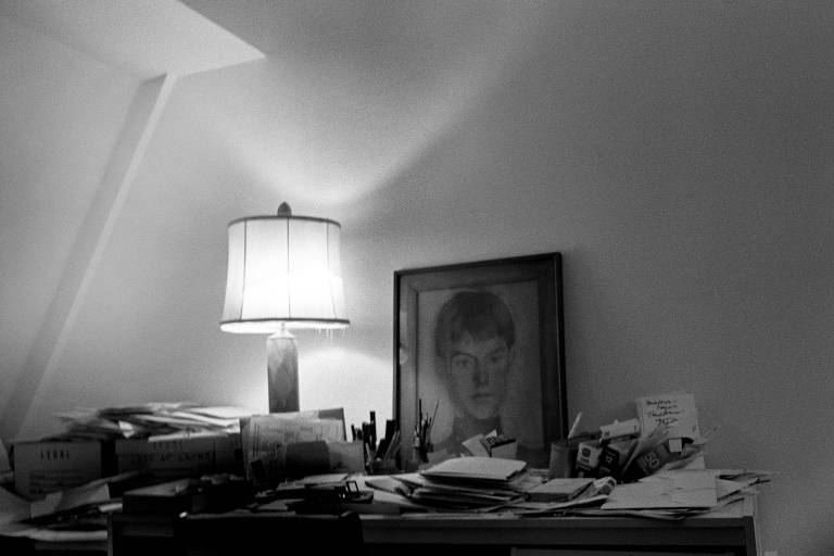 foto em preto e branco de mesa encostada na parede e em cima dela há um abajur aceso e uma pintura de um homem