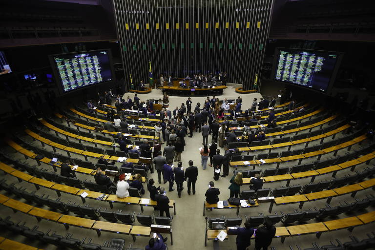 A imagem mostra uma sessão no plenário da Câmara dos Deputados do Brasil. Vários deputados estão reunidos no centro do plenário, enquanto outros estão sentados em suas cadeiras. O ambiente é amplo, com várias fileiras de assentos e mesas. No fundo, há um painel eletrônico grande exibindo informações e resultados de votações.