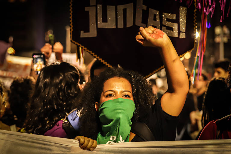 Mulher negra protesta com a mão para o alto e usa um pano verde na boca