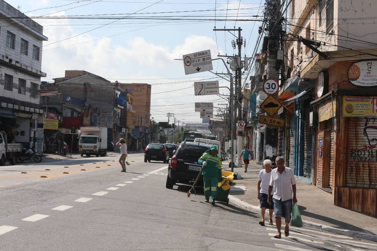 A imagem mostra a avenida Sapopemba durante o dia, com uma faixa de pedestres vazia em primeiro plano e algumas pessoas caminhando na calçada. Há carros estacionados ao lado da via e uma variedade de lojas com fachadas diversas ao longo da rua