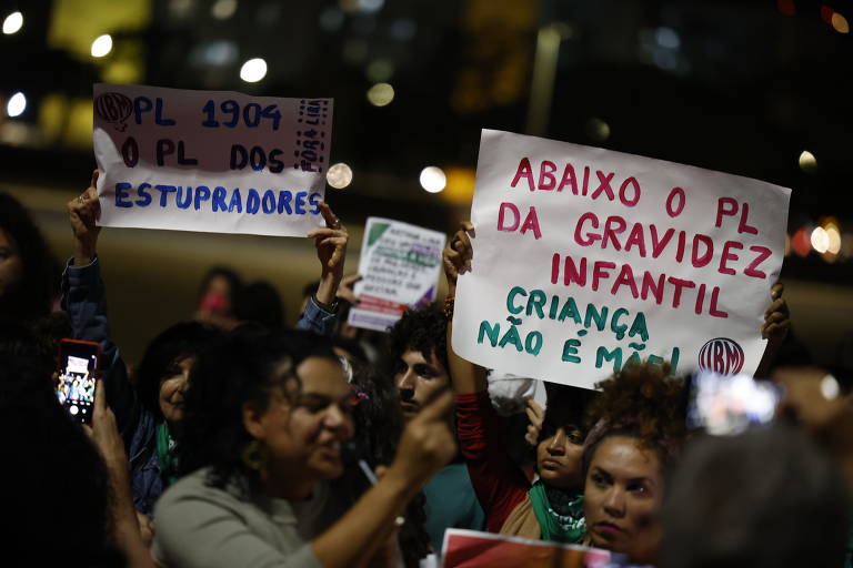 Em um ato de protesto noturno, manifestantes empunham cartazes com mensagens fortes contra o PL antiaborto por estupro