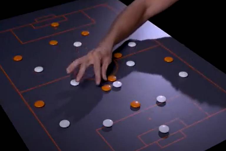 Em uma mesa que imita um campo de futebol e que está repleta de peças laranjas e brancas, a mão direita do treinador Sylvinho mexe em um deles