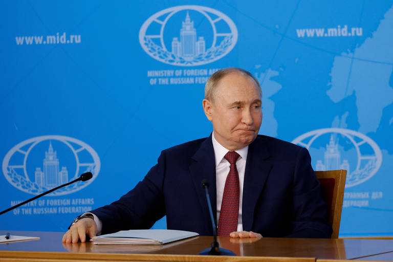 Foto de Vladimir Putin, um homem branco e calvo, usando terno escuro e gravata roxa, em uma mesa com microfones