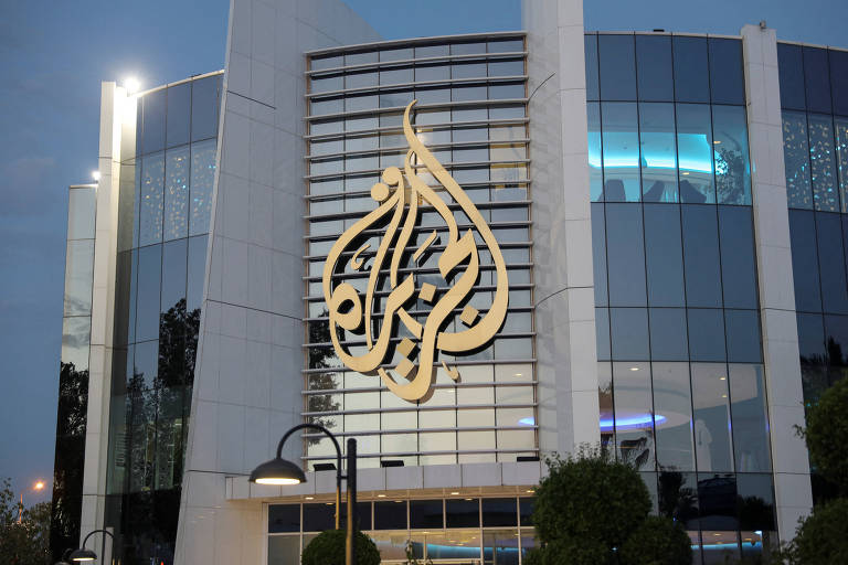 A fachada de um moderno edifício de vidro e aço é iluminada por luzes suaves ao entardecer, destacando o icônico logotipo da Al Jazeera, uma conhecida rede de televisão baseada no Catar, que se projeta em uma elegante caligrafia árabe.