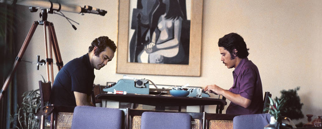 Foto de perfil de dois homens com cabelo escuro sentados frente a frente em uma mesa, cada um trabalhando em uma máquina de escrever. Há um quadro na parede ao fundo e uma luneta à esquerda e, à frente deles, um móvel com estofado azul