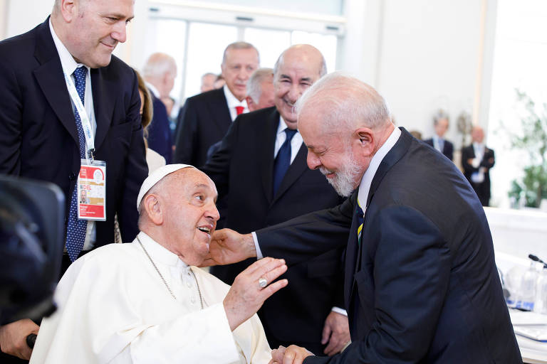 O papa Francisco, vestido com suas vestes papais brancas, compartilha um momento caloroso com o presidente Lula, que se inclina para cumprimentá-lo com um sorriso. Eles parecem estar em um ambiente formal, cercados por outros indivíduos vestidos de maneira formal.