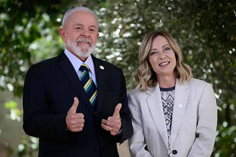 Um homem, Lula, e uma mulher, Giorgia Meloni, ambos vestidos formalmente, estão de pé ao ar livre com um fundo de vegetação. O homem, com barba branca e terno escuro, faz um sinal de positivo com ambas as mãos. A mulher, ao seu lado, veste um blazer claro e sorri.