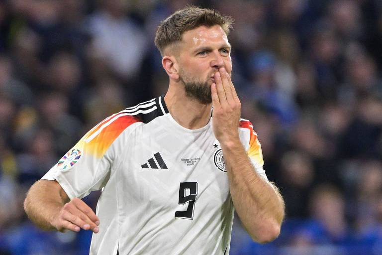 Um jogador de futebol em uniforme branco com detalhes pretos e uma faixa com as cores da bandeira alemã no peito leva a mão à boca em um gesto para celebrar o gol marcado no jogo