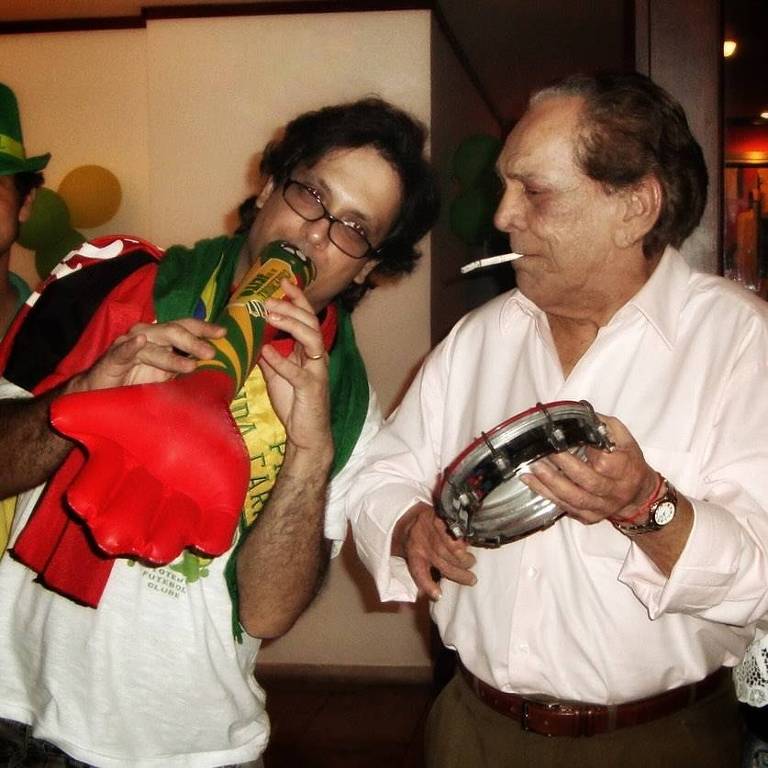 Dois homens, um tocando uma vuvuzela decorada com as cores da bandeira brasileira e o outro um pandeiro