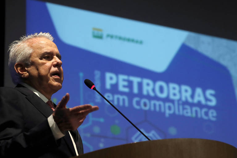 Homem com cabelos grisalhos fala ao microfone em púlpito; ao fundo há uma tela que diz "Petrobras em compliance"