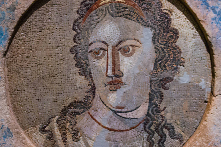 Detalhe de mosaico antigo, representando o rosto de uma figura feminina com traços clássicos. A expressão é serena, com olhos grandes e uma boca delicada, enquadrada por cabelos ondulados e uma coroa ou diadema. O trabalho em mosaico é composto por pequenas peças, criando um efeito visual detalhado e texturizado.