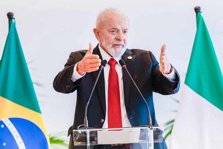 Um homem branco e idoso está em pé, vestido com terno escuro, camisa branca e gravata vermelha. Ele está diante de um púlpito com microfones e com duas bandeiras, um de cada lado dele: à esquerda, a bandeira do Brasil e, à direita, a da Itália