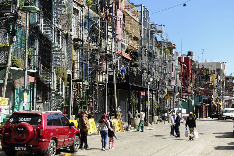 Imagem mostra pessoas andando em rua de uma favela. Casas se amontoam junto com fios em local de baixa renda. Há um carro vermelho estacionado na rua.