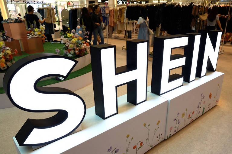 Logo da Shein aparece em primeiro plano. Ao fundo, pessoas circulam em uma loja com vários cabideiros, onde estão dispostas roupas da marca.