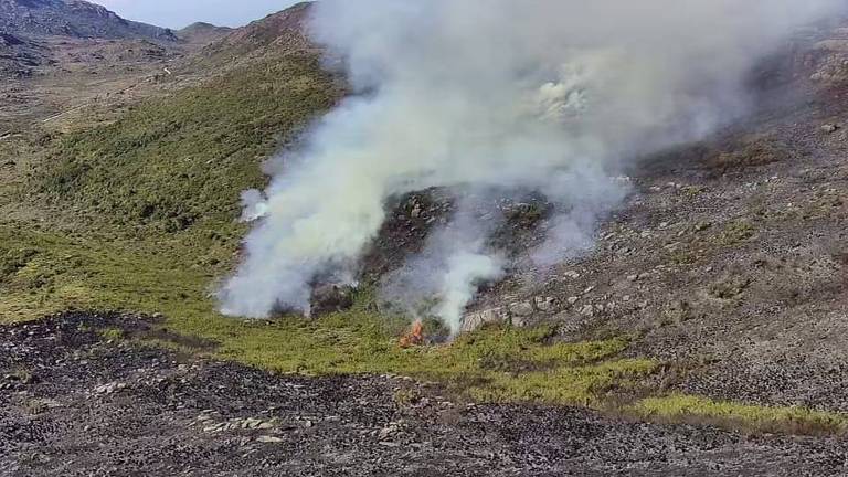 A imagem captura uma paisagem árida e rochosa, onde fumaça branca emerge vigorosamente do solo, devido incêndio que atinge a vegetação. 