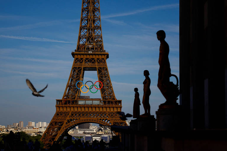 A imagem mostra a Torre Eiffel em Paris, destacando os anéis olímpicos coloridos em sua base. No primeiro plano, há estátuas em silhueta e um pássaro voando. O céu está claro com algumas nuvens.