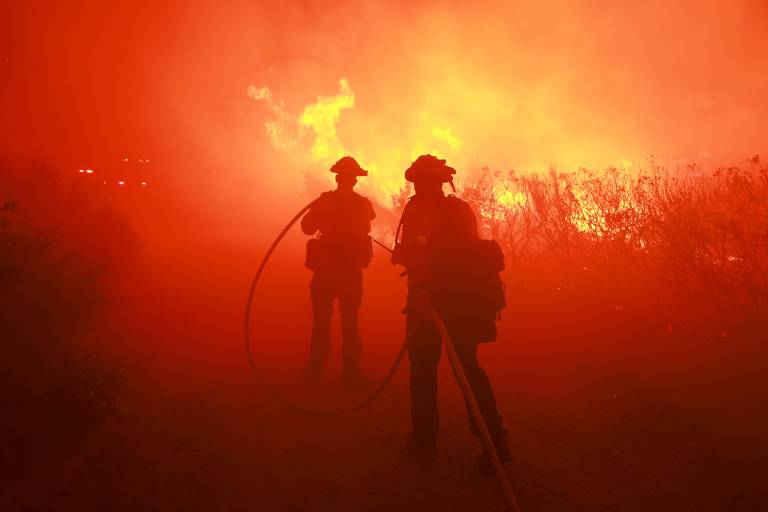 Silhueta de dois bombeiros em meio a muita fumaça e muitas chamas; a foto é completamente vermelha e laranja; há labaredas atrás dos dois homens