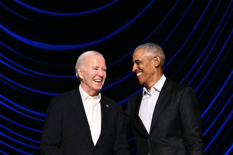 Joe Biden ao lado de Barack Obama. Ambos sorriem e usam terno preto e camisa branca. Ao fundo, listras azuis e espaços pretos em um painel