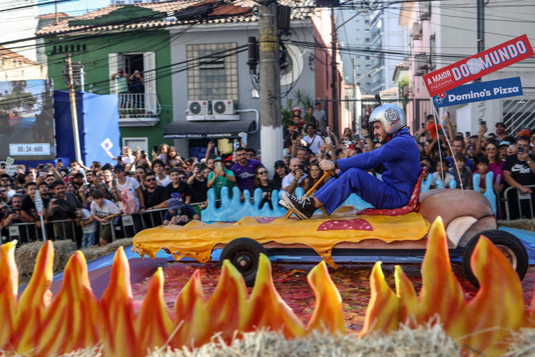 Um participante vestido com um macacão azul e capacete de segurança pilota um carrinho de corrida artesanal que imita uma fatia de pizza gigante. Ele manobra o veículo através de um percurso ladeado por espectadores entusiasmados e decorações que simulam chamas.