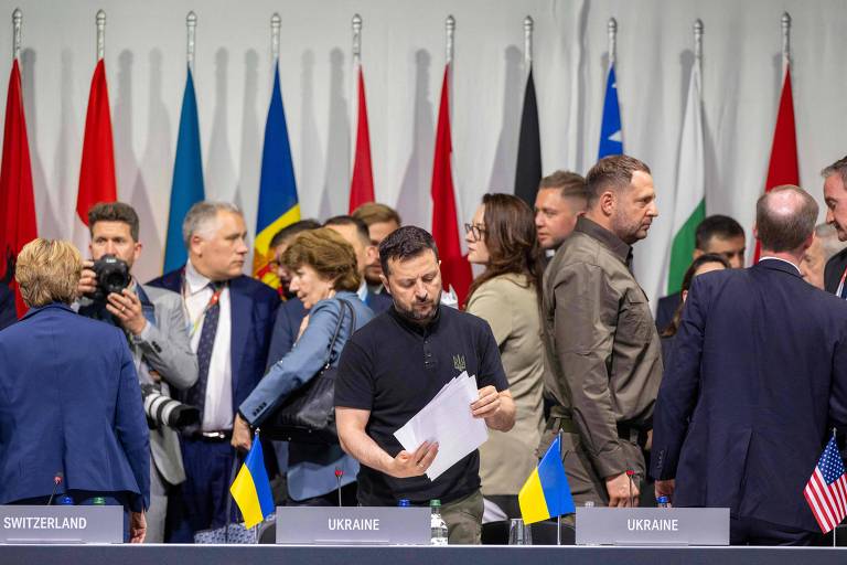 O presidente da Ucrânia, homem de barba e roupa escura, manuseia papel cercado por diversas autoridades num ambiente com bandeiras dos países atrás de uma grande mesa