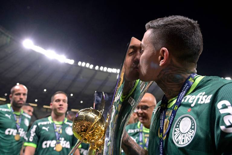 Jogadores de futebol em uniformes verdes comemoram título; um deles beija um troféu dourado. Eles estão em um estádio iluminado, com um deles em primeiro plano, destacando-se pela emoção do momento.
