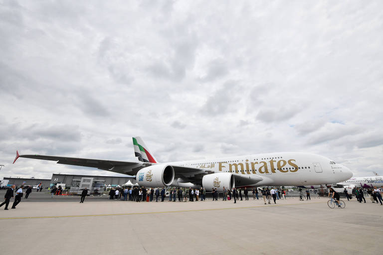 Visitantes se alinham para admirar um imponente avião da Emirates estacionado sob um céu nublado, destacando-se com sua fuselagem branca