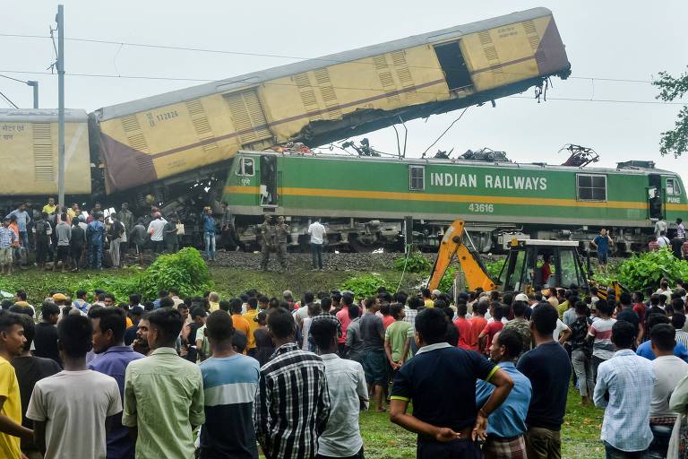 Multidão de espectadores observa o cenário de um acidente ferroviário, onde vagões de carga da Indian Railways estão descarrilados e empilhados de forma caótica. Equipes de emergência trabalham entre os destroços, enquanto uma escavadeira amarela é usada no processo de recuperação.