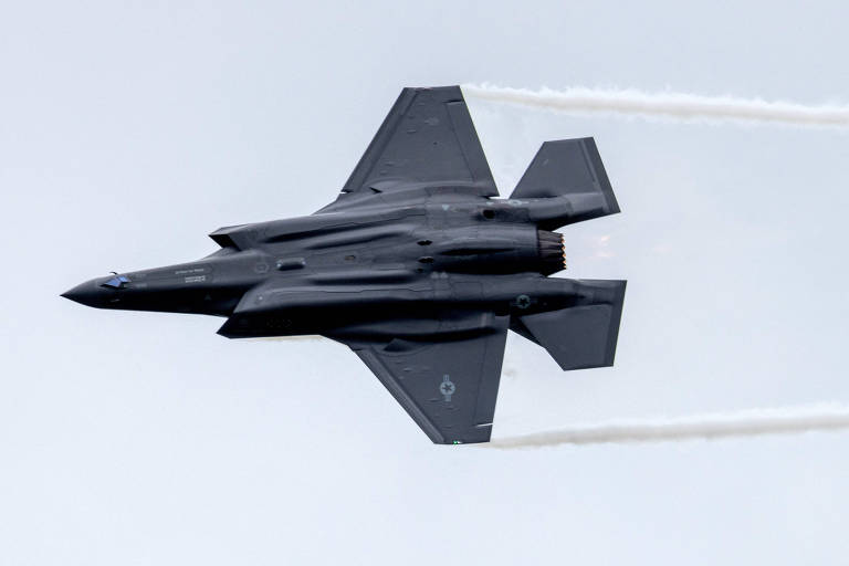 Avião de caça cinza-escuro voa contra céu nublado, com o ventre à mostra e duas trilhas de vapor saindo da ponta de suas asas