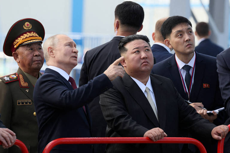 Putin, um homem calvo de terno escuro, aponta algo para Kim, um homem oriental também de terno, observados por um militar uniformizado e um tradutor com caderno na mão