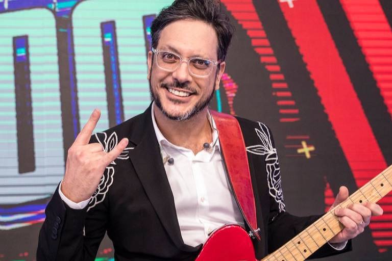 Homem de terno preto e camisa usa óculos enquanto segura uma guitarra e faz um sinal que remete ao rock com a mão direita