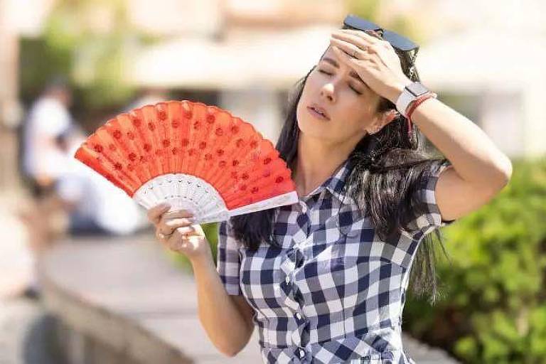 Uma mulher está usando um leque vermelho para se refrescar em um dia ensolarado. Ela veste uma camisa xadrez e usa óculos de sol na cabeça, enquanto parece estar limpando o suor da testa com a outra mão, evidenciando o calor intenso.

