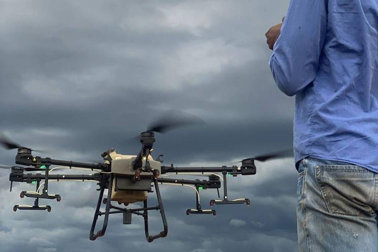 Um homem de camisa azul controla um drone quadricóptero sob um céu nublado.