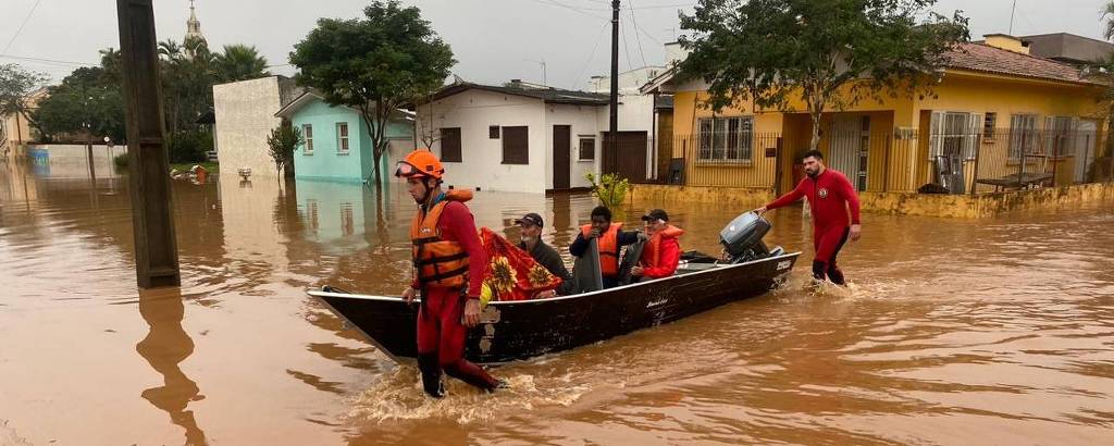Equipe de resgate em trajes laranja navega em um bote por ruas inundadas, passando por casas parcialmente submersas em água barrenta, evidenciando a gravidade da enchente que atingiu a área residencial.
