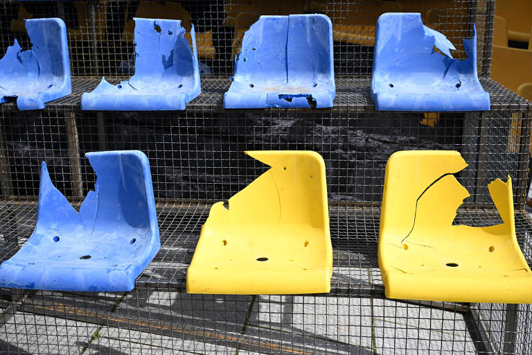 Cadeiras azuis e amarelas quebradas e com marcas de tiros, que estavam em um estadio na Ucrânia