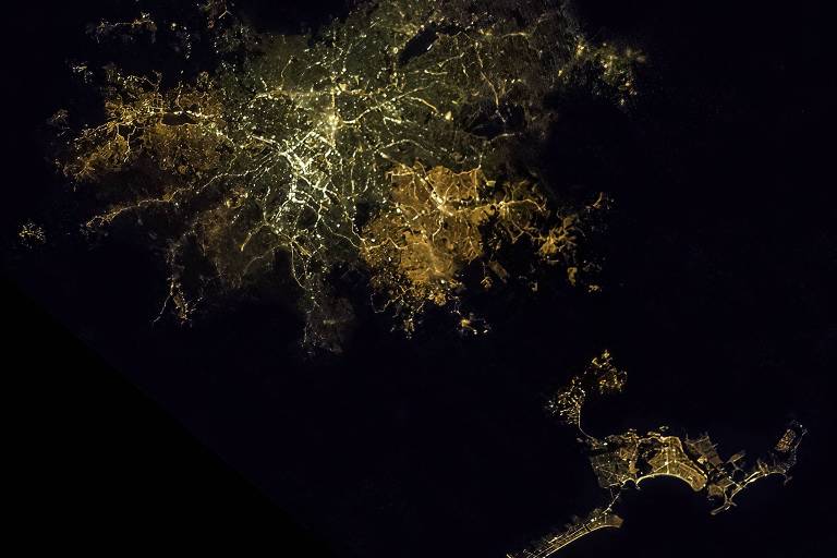 A imagem captura uma vista aérea noturna de uma cidade, com suas luzes formando um padrão complexo e brilhante que contrasta com a escuridão circundante. As luzes amarelas e brancas delineiam as ruas e marcam os centros de atividade, enquanto a escuridão sugere áreas menos povoadas ou naturais.