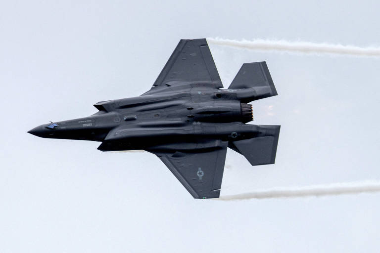 A foto mostra um caça cinza escuro em um voo. Ele está imbicado para a esquerda e uma trilha de fumaça sai de cada asa. Ao fundo, céu nublado.