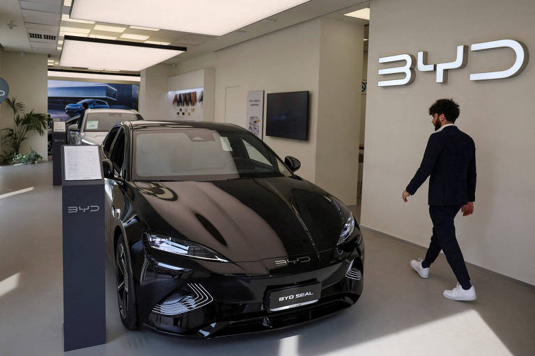 Um homem caminha ao lado de um carro preto em um showroom iluminado, com o logotipo da BYD em destaque ao fundo. 