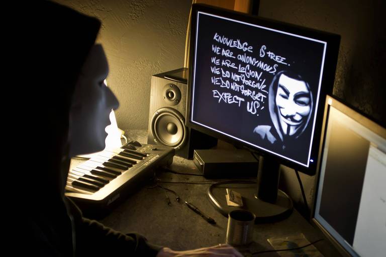 Uma pessoa encapuzada está digitando em um teclado em um ambiente escuro, iluminado apenas pela luz de um monitor que exibe a icônica máscara de Guy Fawkes .