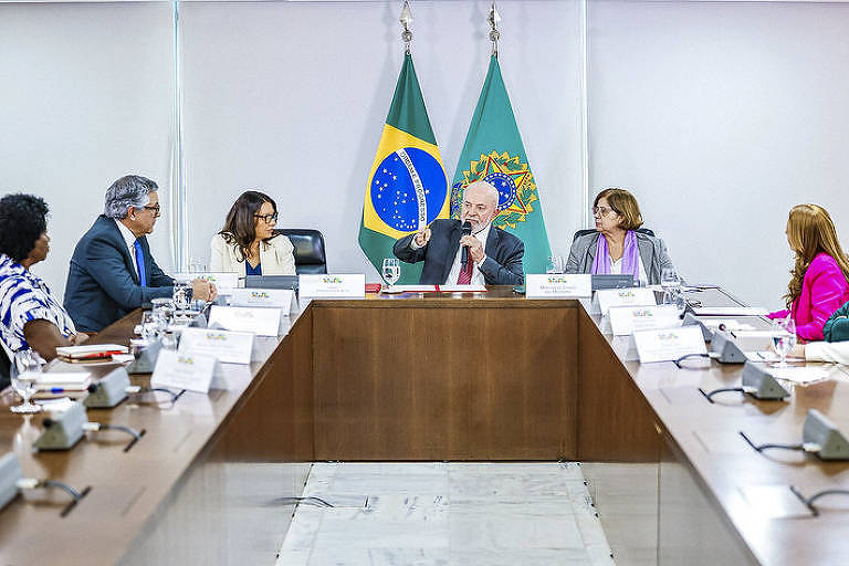 Lula se reúne no Planalto com ministros. Ele está no centro de uma mesa retangular, com bandeiras do Brasil ao fundo. A primeira-dama Janja aparece sentada a seu lado direito, enquanto a ministra das Mulheres, Cida Gonçalves, está do lado esquerdo