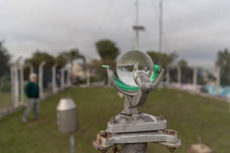 Uma esfera de vidro montada sobre um suporte metálico captura o reflexo invertido do ambiente ao redor, enquanto uma pessoa observa ao fundo, em um dia nublado.