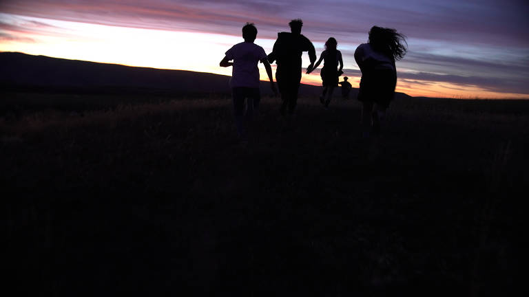 Quatro adolescentes correm em meio a um pôr do sol. Eles estão contra a luz e é possível ver apenas suas silhuetas com o céu alaranjado no fundo