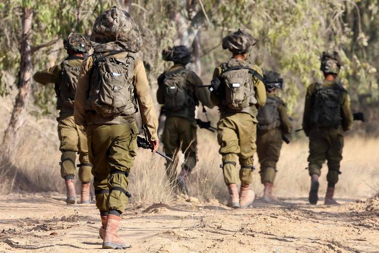 Um grupo de soldados caminha em meio a vegetação seca. Todos estão de costas, vestindo roupas verdes escuras, carregando mochilas e armas.