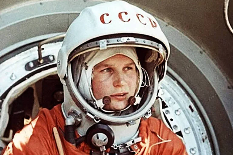 Uma cosmonauta soviética, identificada pelas letras CCCP em seu capacete, aparece em seu traje espacial laranja, pronto para embarcar em uma missão. Seu olhar é concentrado e determinado
