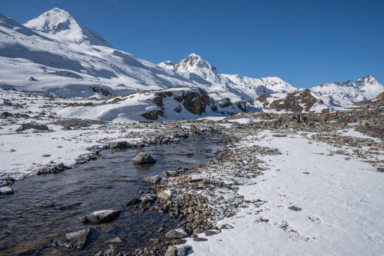 Rio corre em meio a pedras e solo coberto de neve; ao lado há uma montanha coberta de neve